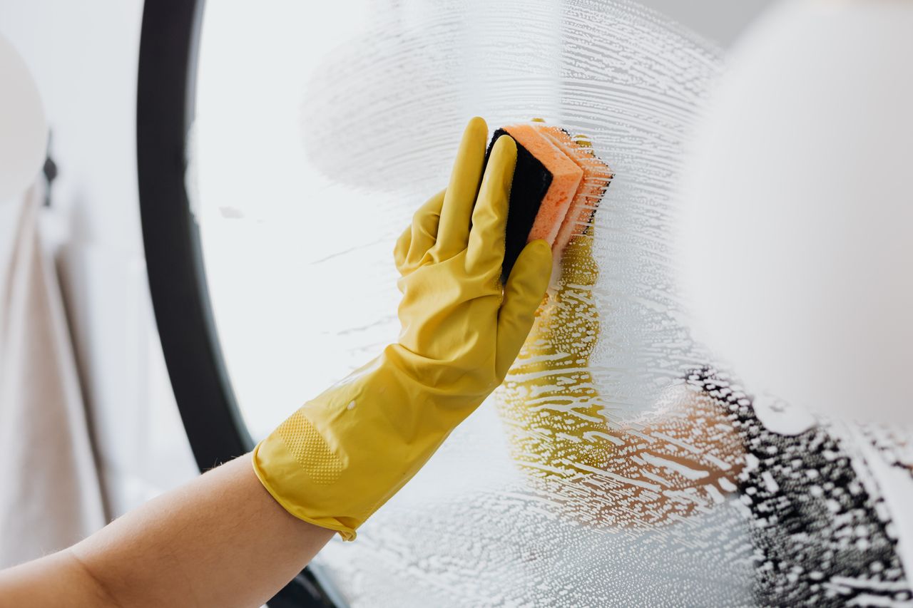 heros-img Anonymous person in yellow rubber gloves using simple sponge for cleaning mirror in bathroom