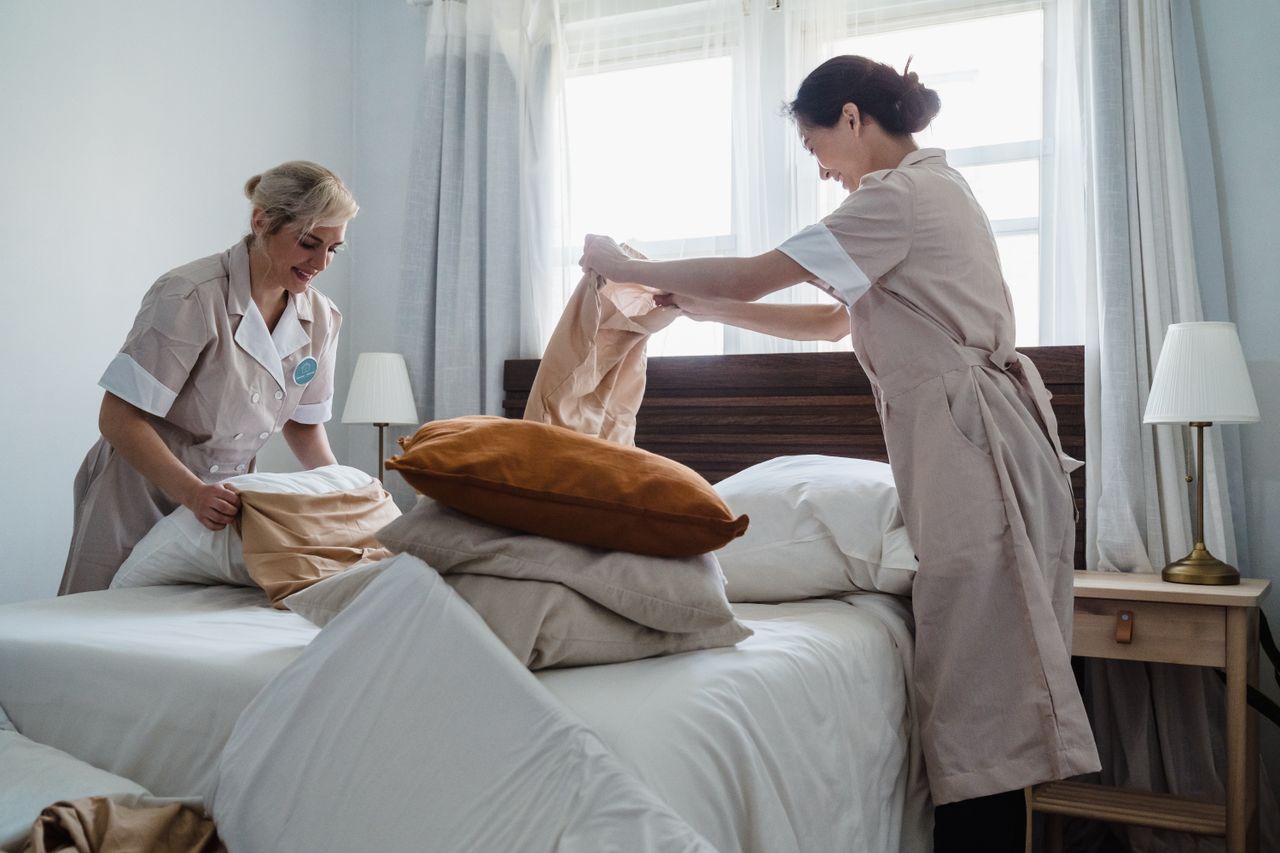 about-us Two housekeepers in uniform fixing a hotel room bed with fresh linen and pillows.